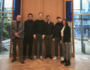 Sechs Personen stehen in einem Innenraum vor einer hohen Holztafel mit blauen Akzenten und großen Fenstern und posieren für ein Gruppenfoto. Die Gruppe besteht aus fünf Männern und einer Frau, die alle leger gekleidet sind. (Text mit KI erstellt)