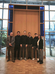 Sechs Personen stehen in einem Innenraum vor einer großen Holzkonstruktion mit blauen vertikalen Paneelen unter einer hohen Glasdecke. Die Gruppe posiert auf einem Parkettboden, hinter ihnen sind durch die Fenster Bäume und Bauwerke zu sehen. (Text mit KI erstellt)
