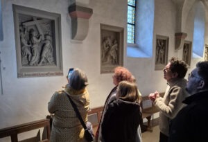 Mehrere Personen stehen in einem Kirchenflur und betrachten und fotografieren religiöse Reliefs an den Wänden unter den Buntglasfenstern. Der Raum ist sanft beleuchtet und hebt die Kunstwerke und die Gruppe hervor. (Text mit KI erstellt)