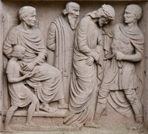 Ein Steinrelief mit der Darstellung von fünf Männern in antiken Gewändern; eine Figur steht zusammengekauert in der Mitte, während die anderen, darunter ein sitzender Mann und ein Kind, um ihn herum stehen und sich zu unterhalten scheinen. (Text mit KI erstellt)