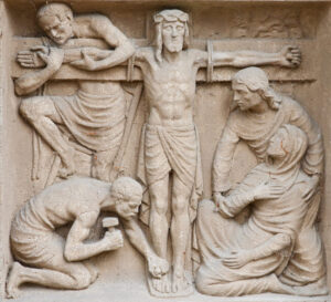 Ein Steinrelief stellt die Kreuzigung Jesu dar, mit Jesus am Kreuz, flankiert von zwei Figuren auf jeder Seite, von denen eine kniet und einen Nagel in seine Füße schlägt, während eine andere Figur eine kniende Frau zu trösten scheint. (Text mit KI erstellt)