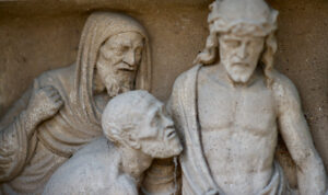 Steinrelief mit der Darstellung dreier bärtiger Männer in Gewändern; eine Figur trägt eine Dornenkrone und blickt nach unten, während die anderen eng mit ihm zu kommunizieren scheinen. Die Szene deutet auf einen Moment der Trauer oder der Ehrfurcht hin. (Text mit KI erstellt)