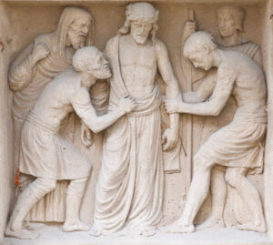 Flachreliefskulptur von fünf Männern; die zentrale Figur wirkt traurig und trägt eine Dornenkrone, während zwei Männer zu beiden Seiten an seinem Gewand ziehen und zwei weitere hinter ihnen stehen. (Text mit KI erstellt)