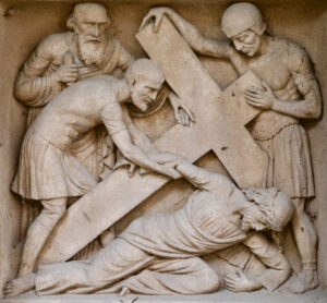 Ein Steinrelief stellt vier Männer dar; einer ist unter einem großen Kreuz auf den Boden gefallen, während zwei Männer ihm beistehen und ein weiterer feierlich zuschaut. Die Szene scheint von den Stationen des Kreuzwegs zu stammen. (Text mit KI erstellt)