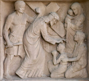 Ein Steinrelief zeigt Jesus, der ein Kreuz trägt und ein kniendes Kind segnet, während zwei Frauen - eine hält ein Baby - neben dem Kind knien. Hinter Jesus steht ein Wächter, der einen Stock hält. (Text mit KI erstellt)