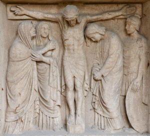 Steinrelief mit der Darstellung der Kreuzigung Jesu, mit Jesus am Kreuz, flankiert von trauernden Figuren und einem römischen Soldaten, der einen Schild hält. (Text mit KI erstellt)