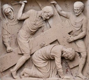 Ein Steinrelief zeigt drei Männer: Einer kniet und trägt ein großes Kreuz, während die beiden anderen stehen, einer hält eine Peitsche, die ihn vorwärts zu zwingen scheint. Die Szene vermittelt den Eindruck von Kampf und Last. (Text mit KI erstellt)