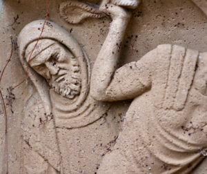 Nahaufnahme eines verwitterten Steinreliefs, das eine bärtige Figur in einem Kapuzengewand darstellt, die einen Arm hochhält. Die Oberfläche weist Altersspuren auf, mit kleinen Rissen und Pflanzenranken. (Text mit KI erstellt)