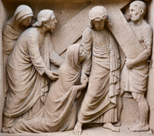 Ein Steinrelief zeigt vier Figuren, die eine kniende Frau trösten, während ein Mann, der ein Gewand und eine Dornenkrone trägt, ein großes Holzkreuz hält. Die Szene ist mit ausdrucksstarken Gesten und drapierter Kleidung detailliert dargestellt. (Text mit KI erstellt)