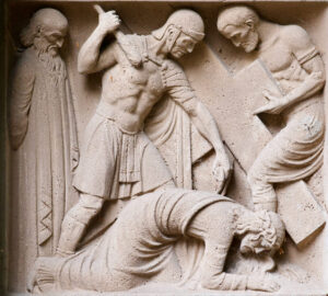 Ein Steinrelief zeigt einen Soldaten mit erhobenem Schwert, einen anderen Mann mit einem Buch in der Hand und eine kniende Figur mit gesenktem Kopf, während ein älterer bärtiger Mann an der Seite steht; alle tragen antike Kleidung. (Text mit KI erstellt)