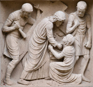 Das Steinrelief zeigt vier Männer in Gewändern; eine zentrale Figur steht und berührt den Kopf eines knienden Mannes, während zwei andere in der Nähe stehen und die Szene beobachten. Der Stil ist klassisch und in hellem Stein gemeißelt. (Text mit KI erstellt)