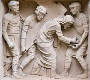 Ein Steinrelief stellt vier Männer dar; die zentrale Figur, die ein Gewand und eine Dornenkrone trägt, trägt ein großes Kreuz, während zwei Männer ihm beistehen und ein vierter zusieht, alle in altrömischer Kleidung. (Text mit KI erstellt)