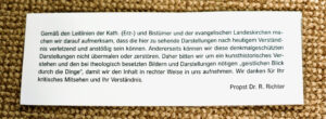 Ein gedruckter deutscher Hinweis auf weißem Papier, der erklärt, dass einige historische religiöse Bilder heute als anstößig empfunden werden können, und die Betrachter um Verständnis bittet; er ist von Propst Dr. R. Richter unterzeichnet und auf einer strukturierten Oberfläche angebracht. (Text mit KI erstellt)