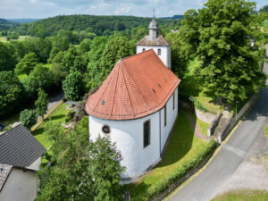 Luftaufnahme einer kleinen weißen Kirche mit rotem Ziegeldach und Glockenturm, umgeben von üppigen grünen Bäumen und Feldern, neben einer ruhigen Straße in einer ländlichen Gegend. (Text mit KI erstellt)