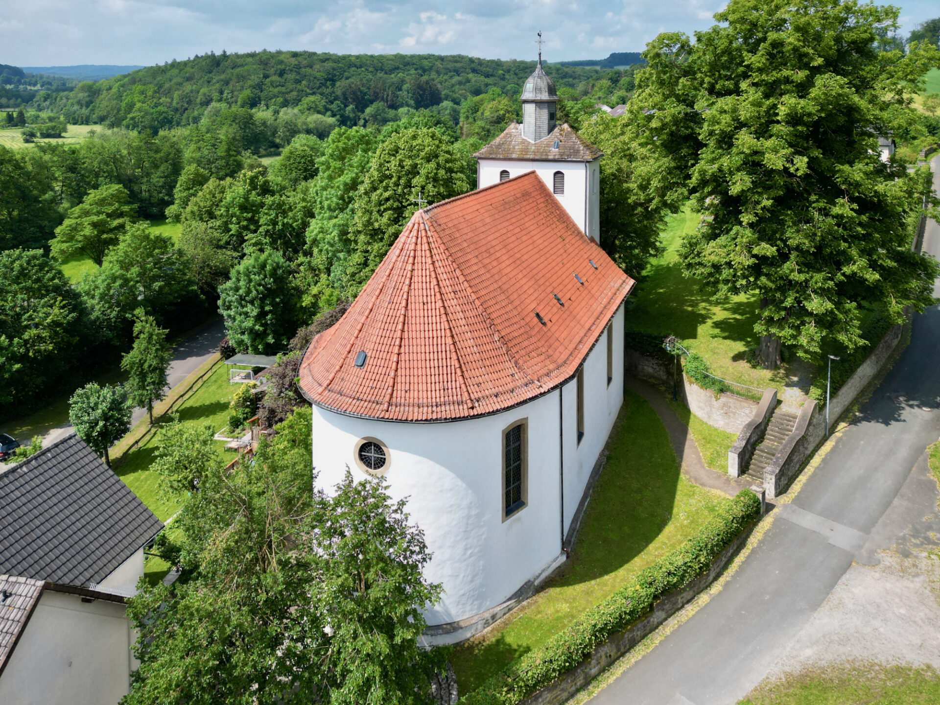 Luftaufnahme einer kleinen weißen Kirche mit rotem Ziegeldach und Glockenturm, umgeben von üppigen grünen Bäumen und Feldern, neben einer ruhigen Straße in einer ländlichen Gegend. ( )