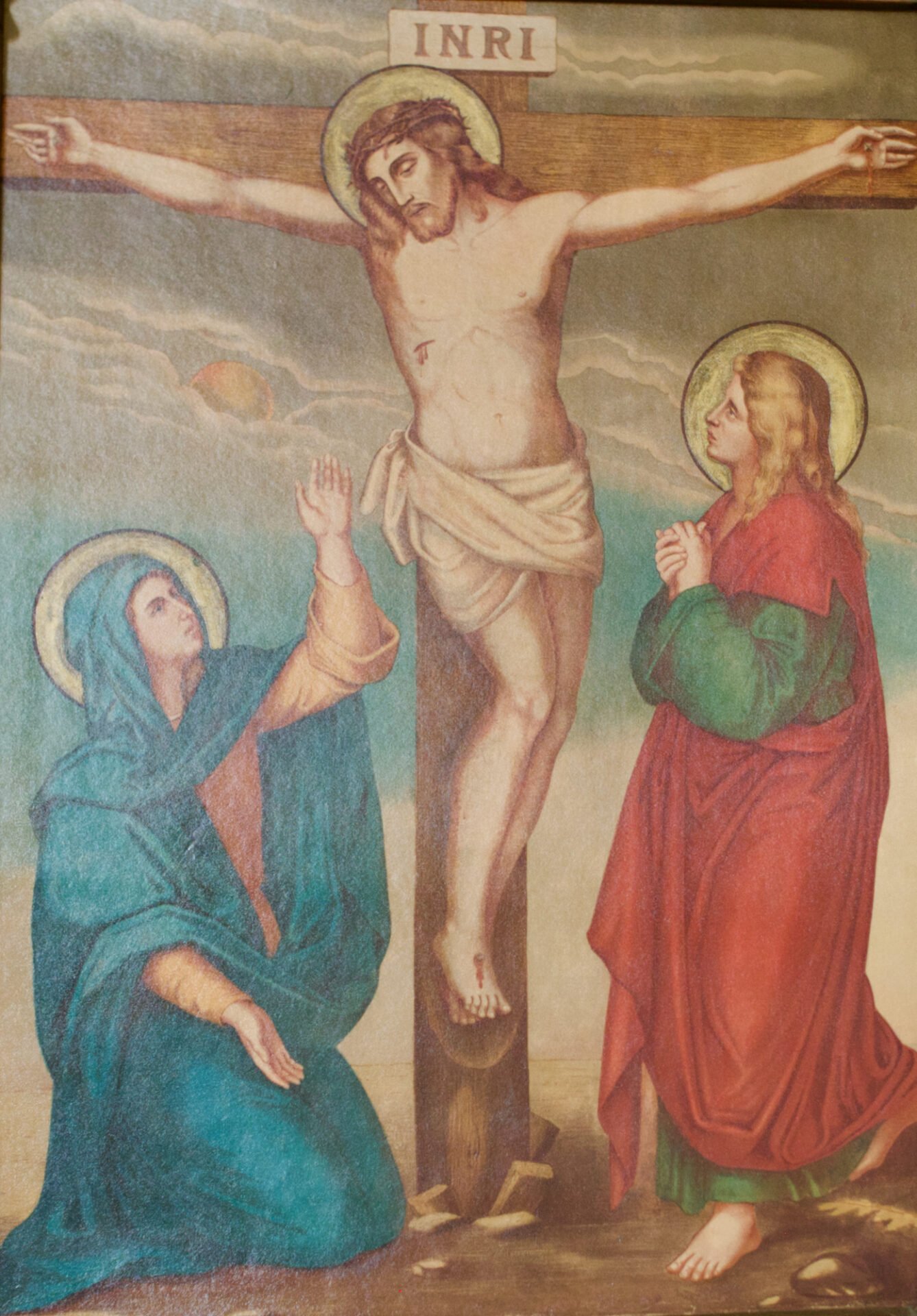 Ein Gemälde von Jesus am Kreuz mit dem Zeichen INRI über ihm, Maria in Blau, die kniet und sich aufrichtet, und eine weitere Figur in Rot und Grün, die neben dem Kreuz steht und betet. ( )