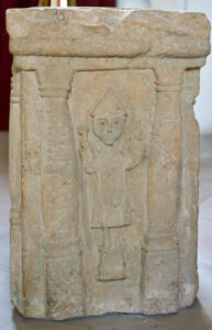 Geschnitztes Steinrelief, das eine stehende Figur mit Spitzhut und einem Gegenstand in der Hand darstellt, eingerahmt von zwei Säulen und einem Bogen; der Stein hat eine verwitterte Oberfläche. (Text mit KI erstellt)