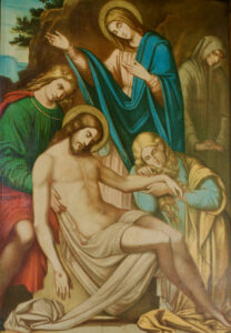 Ein klassisches Gemälde zeigt den leblosen Körper Jesu, der von Trauernden gewiegt wird, darunter eine trauernde Frau in Blau, eine weitere in Gelb, die seine Hand hält, und zwei weitere trauernde Gestalten, alle vor einem gedämpften Hintergrund. (Text mit KI erstellt)
