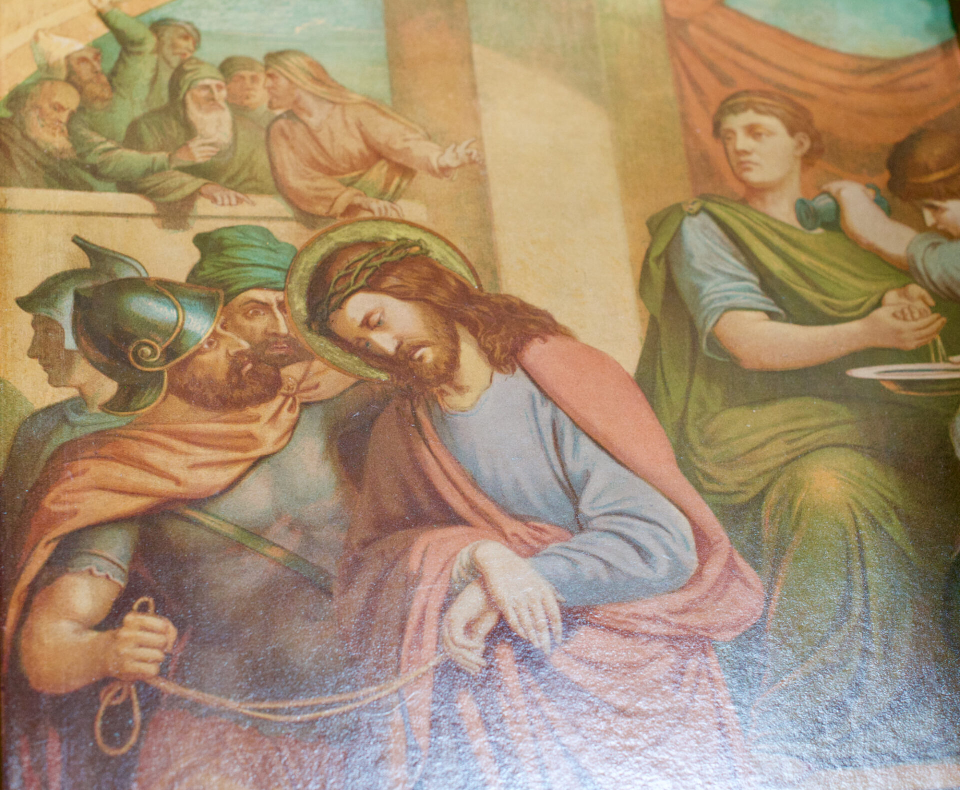 Ein Gemälde zeigt Jesus mit einer Dornenkrone, umgeben von römischen Soldaten und Schaulustigen. Ein Soldat hält ein Seil, mit dem die Hände Jesu gefesselt sind, während ein Mann in Grün an einem Tisch im Hintergrund seine Hände zu waschen scheint. ( )