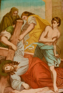 Ein Gemälde zeigt Jesus, der unter dem Gewicht eines Holzkreuzes zusammengebrochen ist, umgeben von drei Männern, von denen einer eine Peitsche hält und ein anderer das Kreuz trägt. (Text mit KI erstellt)