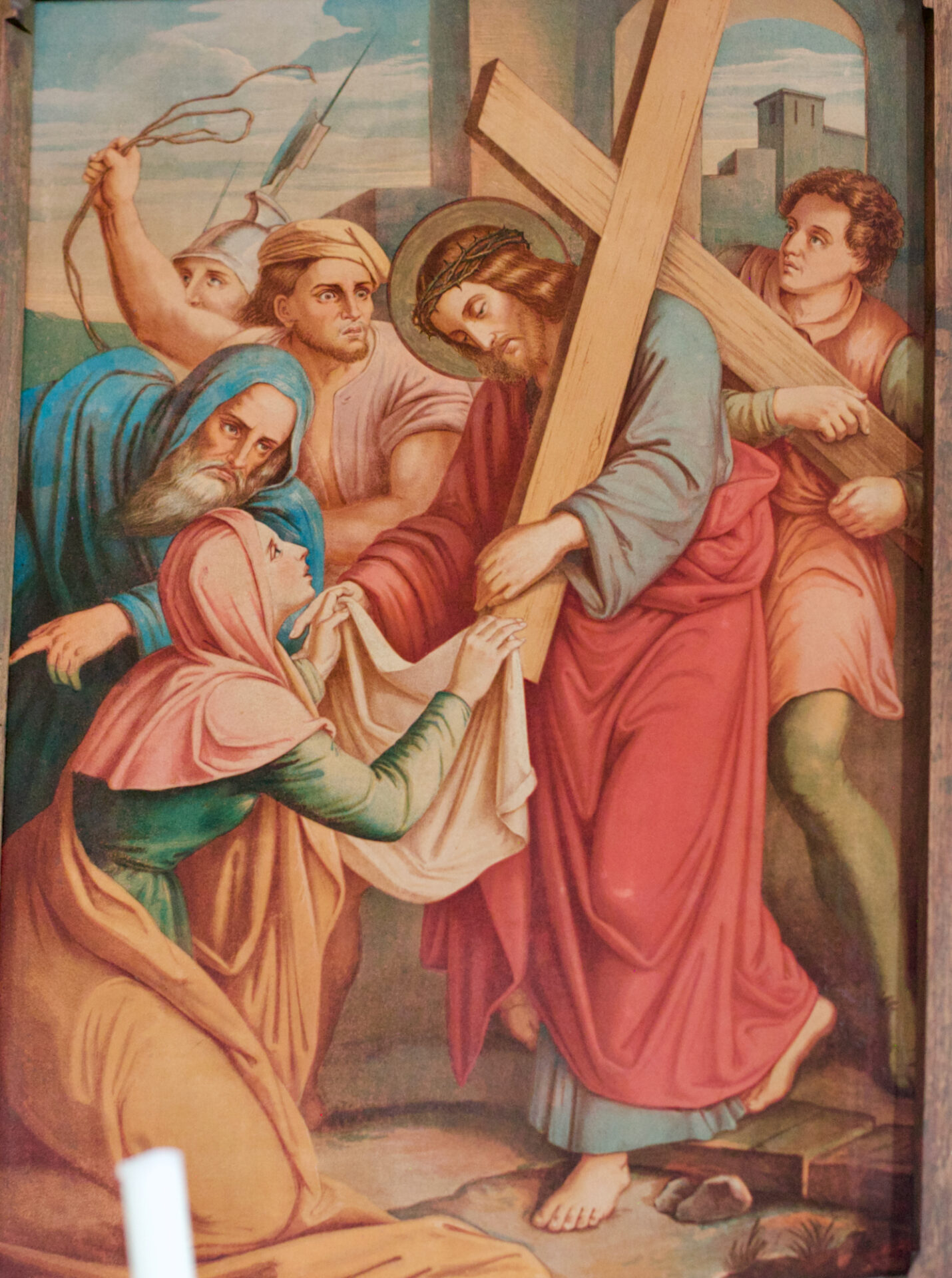 Ein Gemälde zeigt Jesus, der ein Holzkreuz trägt, umgeben von mehreren Figuren. Eine kniende Frau hält ihm ein Tuch vor das Gesicht, während andere besorgt zusehen. Im Hintergrund sind Gebäude und ein bewölkter Himmel zu sehen. ( )