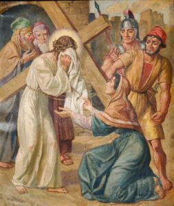 Eine Frau kniet nieder und bietet einem Mann, der ein Kreuz trägt, ein Tuch an. Sie ist umgeben von Schaulustigen und römischen Soldaten und stellt die Passion Christi dar. (Text mit KI erstellt)