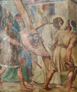 Ein Gemälde zeigt Jesus mit einem Holzkreuz und einer Dornenkrone, umgeben von römischen Soldaten und einem Mann, der ihm hilft, während ein anderer Mann sie mit einer Peitsche führt. Im Hintergrund sind einfache Gebäude zu sehen. (Text mit KI erstellt)