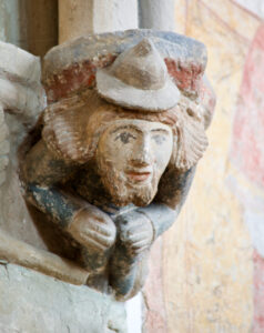 Eine in Stein gemeißelte bärtige Figur mit Hut, die in verblassten Farben gemalt ist, lehnt mit gefalteten Händen vor einer alten, verwitterten Wand mit Spuren von Wandmalerei. (Text mit KI erstellt)