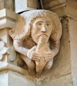 Eine in Stein gemeißelte Figur mit langem Haar, die in der Hocke sitzt und scheinbar ihren eigenen Fuß in den Mund nimmt, inmitten architektonischer Details eines alten Gebäudes. (Text mit KI erstellt)