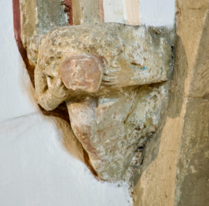 Eine verwitterte Steinskulptur eines menschlichen Gesichts und menschlicher Hände, die wie aus einer Mauerecke herausragt, mit sichtbarer Erosion und verblassten Details. Die umgebende Wand ist weiß mit einigen freigelegten Steinen. (Text mit KI erstellt)