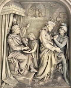 Ein detailliertes Steinrelief mit einer sitzenden Figur, die sich die Hände wäscht, einem Kind, das eine Schale hält, und zwei stehenden Figuren - eine davon in Ketten - neben einem Wächter, eingefasst in einen kunstvollen architektonischen Rahmen. (Text mit KI erstellt)