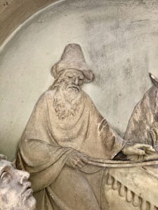 Eine Steinreliefskulptur, die einen älteren bärtigen Mann mit hohem Hut und Gewand zeigt, der sich mit düsterer Miene an etwas festhält, das ein drapiertes Objekt oder Fahrzeug zu sein scheint. Der Hintergrund ist strukturiert und sanft geschnitzt. (Text mit KI erstellt)
