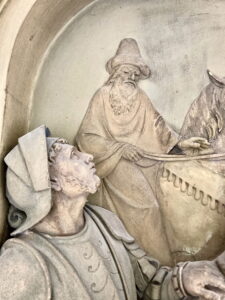 Die Nahaufnahme einer Steinreliefskulptur zeigt zwei Männer in historischer Kleidung; der eine trägt einen Hut und hält beim Reiten die Zügel, der andere, in Rüstung, blickt mit besorgtem Blick nach oben. (Text mit KI erstellt)