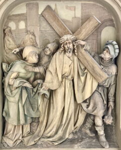 Eine steinerne Reliefskulptur zeigt Jesus, der ein großes Holzkreuz trägt, umgeben von mehreren Figuren in Gewändern und Rüstungen, mit Gebäuden im Hintergrund, die eine Szene des Kreuzweges darstellen. (Text mit KI erstellt)