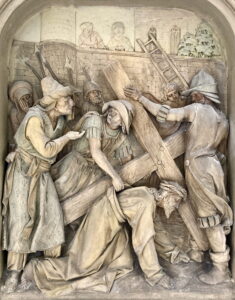 Ein detailliertes Steinrelief zeigt mehrere Figuren in historischer Kleidung, die sich bemühen, ein großes Holzkreuz zu tragen. Ein älterer Mann beugt sich darunter, während andere ihm helfen oder ihn anspornen, vor dem Hintergrund einer Mauer und von Leitern. (Text mit KI erstellt)