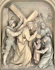 Ein detailliertes Steinrelief stellt Jesus dar, der ein großes Holzkreuz trägt und von Soldaten und Trauernden umgeben ist. Ein Kind und zwei Frauen reichen ihm die Hand, während ein Wächter mit einer Hand gestikuliert und Umstehende von einem Balkon im Hintergrund zusehen. (Text mit KI erstellt)