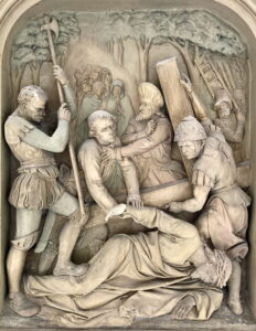 Ein Steinrelief, das mehrere gepanzerte Figuren zeigt, die einen Mann mit einem großen Holzkreuz zu Boden zwingen, während andere hinter ihnen stehen und Bäume im Hintergrund zu sehen sind. (Text mit KI erstellt)