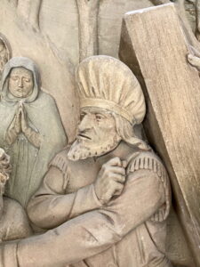 Ein Steinrelief zeigt einen bärtigen Mann mit hohem Hut und Gewand, der sich mit ernster Miene an einem großen rechteckigen Gegenstand festhält. Hinter ihm betet eine Frau mit bedecktem Kopf und gefalteten Händen. (Text mit KI erstellt)