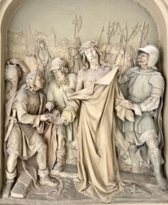 Ein detailliertes Steinrelief zeigt Jesus mit einer Dornenkrone, umgeben von römischen Soldaten und Anhängern. Ein Mann kniet und bietet Jesus ein Gewand an, während andere im Hintergrund Speere und Schilde halten. (Text mit KI erstellt)