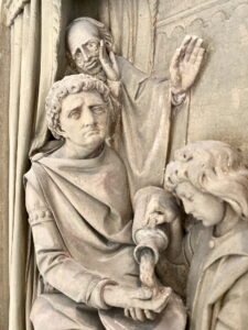 Ein detailliertes Steinrelief zeigt drei Figuren: Eine gießt Wasser in die Hand einer anderen, die feierlich dreinschaut, während eine dritte Figur im Hintergrund eine Hand hebt und nachdenklich blickt. Die Szene vermittelt den Eindruck eines Rituals oder einer Zeremonie. (Text mit KI erstellt)