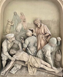 Eine detaillierte Reliefskulptur stellt mehrere Figuren dar, die den Leichnam Jesu nach der Kreuzigung umgeben. Einige Figuren trösten sich gegenseitig, während andere den leblosen Körper anfassen und so eine Szene der Trauer und des Mitgefühls schaffen. (Text mit KI erstellt)