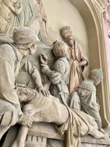 Steinrelief mit mehreren Figuren, die sich um einen leblosen, horizontal aufgebahrten Mann kümmern, mit feierlichem Gesichtsausdruck und detaillierter zeitgenössischer Kleidung, vor einem architektonischen Hintergrund. (Text mit KI erstellt)