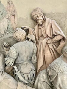 Ein detailliertes Steinrelief zeigt einen bärtigen Mann in einem Gewand, der auf eine kniende gepanzerte Figur mit einem Schwert zeigt. Um sie herum sind teilweise weitere Personen zu sehen, was auf eine historische oder biblische Szene schließen lässt. (Text mit KI erstellt)