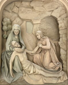 Eine Reliefskulptur zeigt zwei Frauen, die um den Leichnam eines Mannes, wahrscheinlich Jesus, trauern, in einer Szene, die an die Pietà erinnert. Der Hintergrund zeigt eine felsige Höhle oder den Eingang eines Grabes. Die Figuren sind in Gewänder gehüllt und wirken feierlich. (Text mit KI erstellt)