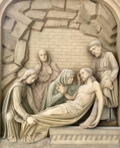 Ein Steinrelief zeigt den Leichnam Jesu, der von vier Figuren, zwei Frauen und zwei Männern, in einem höhlenartigen Grab mit einer Ziegelwand im Hintergrund beweint wird. (Text mit KI erstellt)