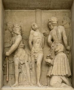 Eine Steinreliefskulptur zeigt eine gefesselte Figur in der Mitte, flankiert von zwei Männern mit Peitschen und einem Mann in kunstvoller Kleidung, der kniet und einen Gegenstand hält, alles vor einem detaillierten Hintergrund. (Text mit KI erstellt)