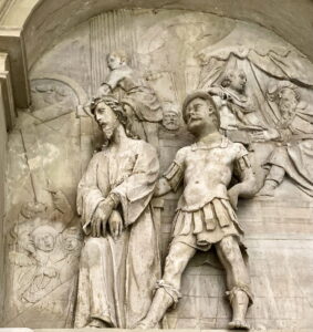 Steinrelief, das eine in ein Gewand gehüllte Figur mit gefesselten Händen zeigt, die neben einem römischen Soldaten steht; im Hintergrund schauen geschnitzte Figuren hinter drapierten Vorhängen hervor, und teilweise sind Engelsgesichter zu sehen. (Text mit KI erstellt)