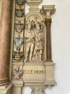 Steinrelief mit drei Figuren, eine davon dornengekrönt, umgeben von kunstvollen architektonischen Details und heraldischen Schilden, darunter die Inschrift "Ecce Homo". (Text mit KI erstellt)