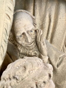 Nahaufnahme einer verwitterten Steinskulptur, die einen bärtigen Mann mit sanftem Gesichtsausdruck zeigt, der seine Wange auf die Hand stützt, mit komplizierten Details in Gesicht und Gewand. (Text mit KI erstellt)