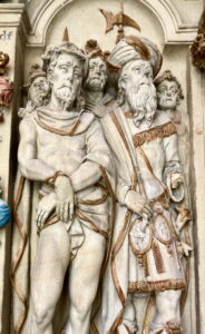 Ein detailliertes Steinrelief, das in der Mitte zwei männliche Figuren in antiker Kleidung zeigt, eine mit Dornenkrone und gefesselten Händen, die andere mit Turban und Gewand, umgeben von Soldaten mit Helmen und ernster Miene. (Text mit KI erstellt)